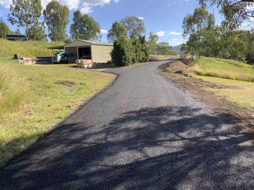 Freshly laid bitumen driveway leading to a rural shed, surrounded by lush greenery and trees under a clear blue sky.