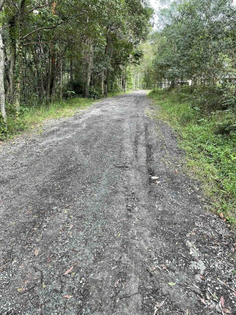 Unstable driveway surface with poor drainage and loose gravel over organic soil, showing signs of erosion and inadequate base preparation.